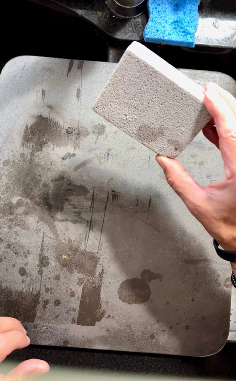 Hand holding a pumice cleaning brick over a well-used Baking Steel in the sink, ready to scrub