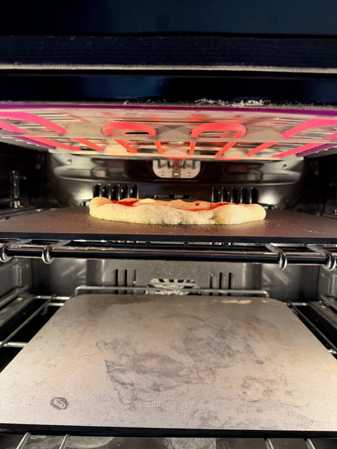 Two Baking Steel Originals stacked in a home oven, with a pizza cooking on the top steel under a glowing broiler — the setup for making multiple pizzas fast.