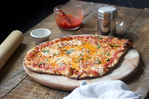 Homemade pizza with charred crust and fresh basil on a wooden pizza peel with crushed tomato sauce and rolling pin in the background