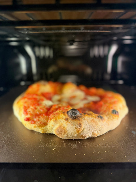 Margherita pizza with a puffy, blistered, char-spotted crust baking on a Baking Steel inside a home oven, brand name embossed on the steel surface