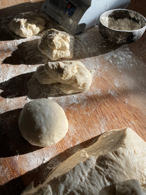 Pizza dough balls in various stages of shaping on a floured wood board with a kitchen scale