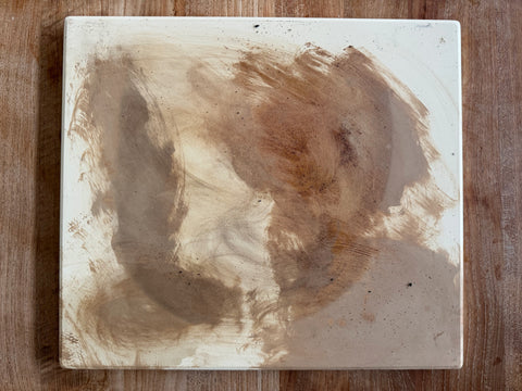 A worn ceramic pizza stone on a wood counter, its surface stained with dark brown grease marks and specks from years of absorbed oils — the buildup that causes stones to smell rancid over time.
