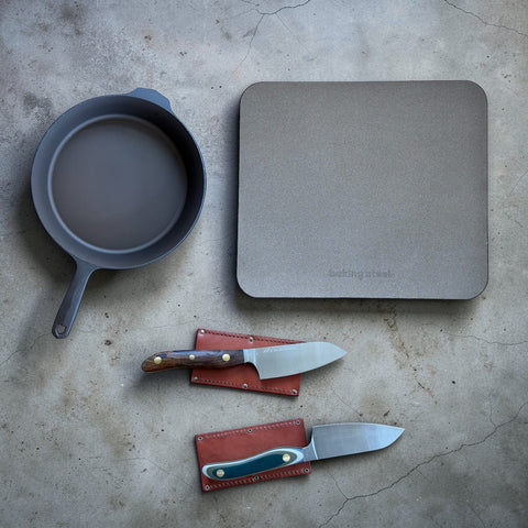 Baking Steel Original, Field Company cast iron skillet, and New West KnifeWorks chef knives with leather sheaths on concrete surface